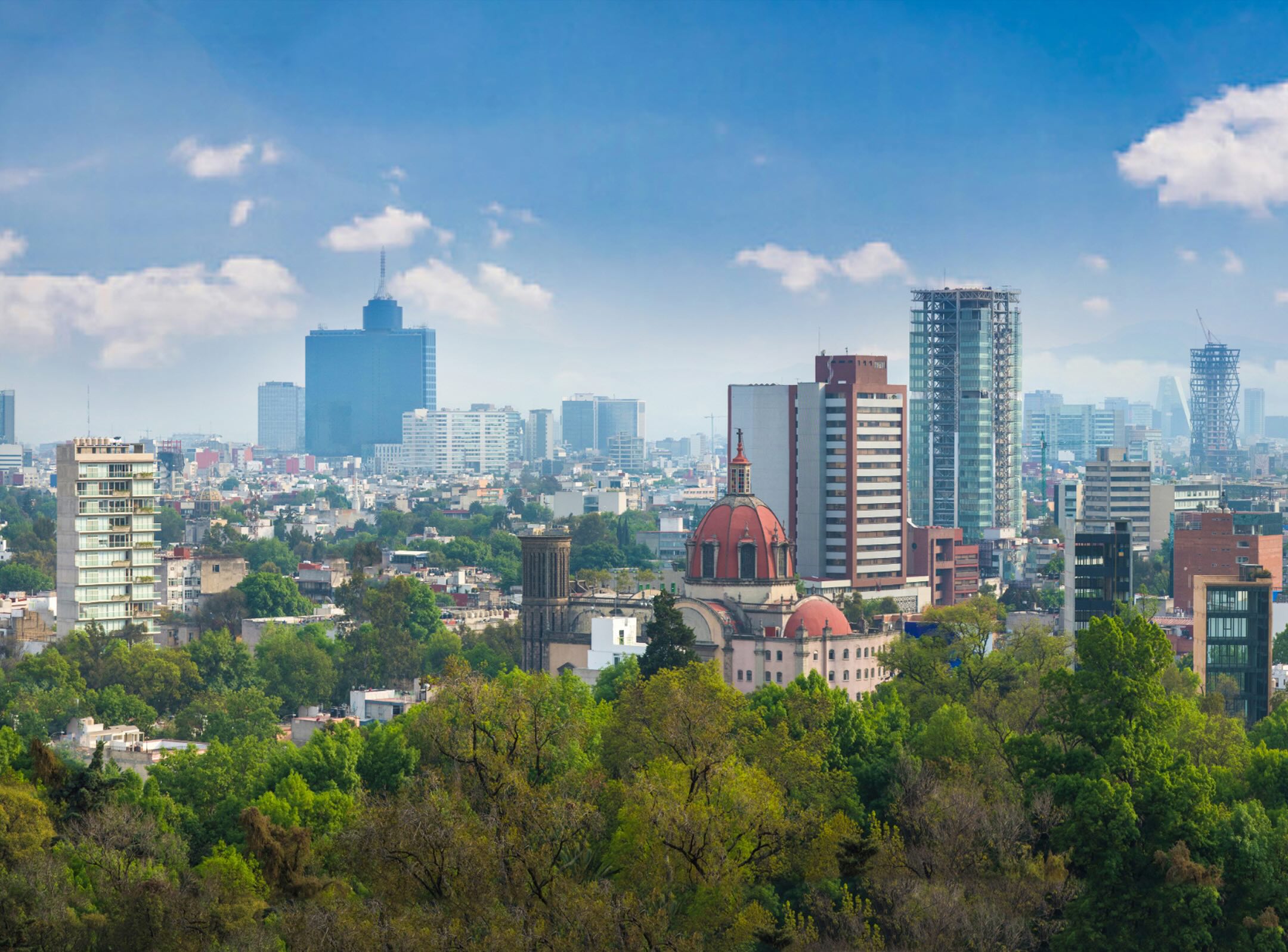 City skyline with tall modern buildings, a historic domed church, and lush green trees in the foreground under a blue sky with scattered clouds.