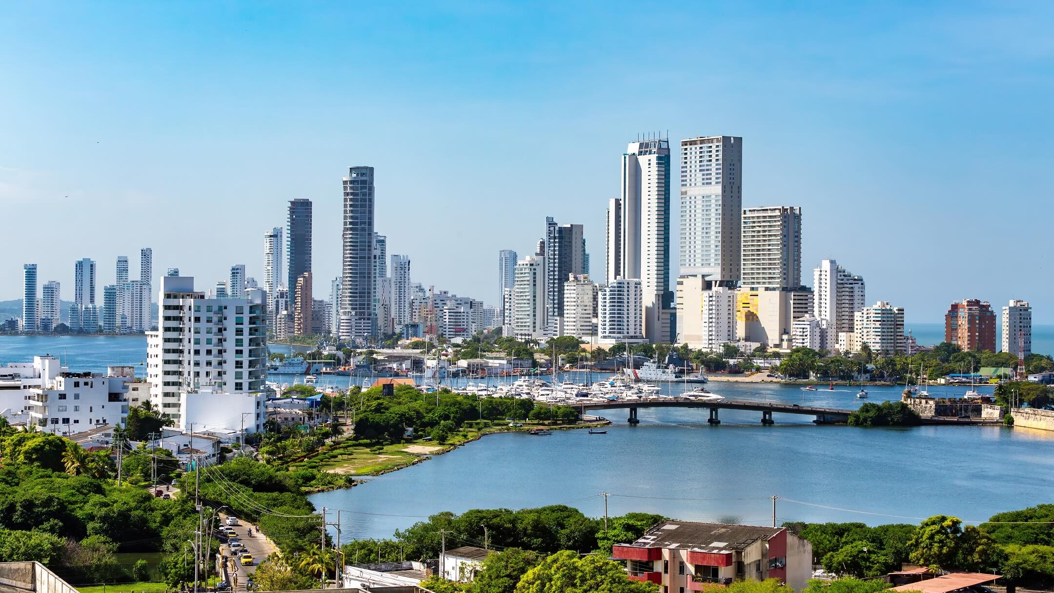 Skyline of a coastal city with tall modern buildings, a marina with boats, a blue river, green trees in the foreground, and a clear blue sky.