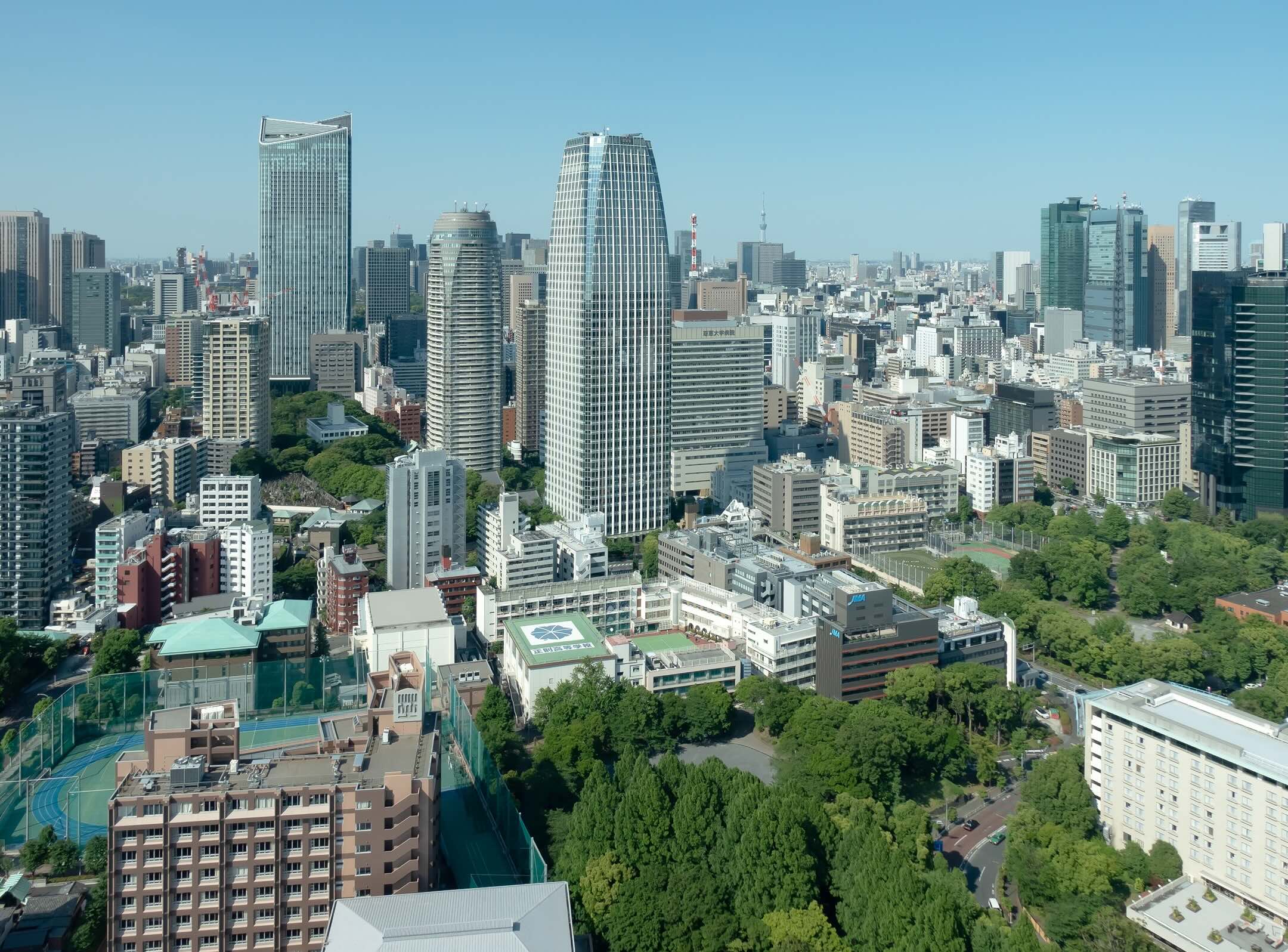 A cityscape featuring tall modern skyscrapers, mid-rise buildings, and green spaces under a clear blue sky. The scene shows a dense urban area with diverse architecture and tree-lined areas.