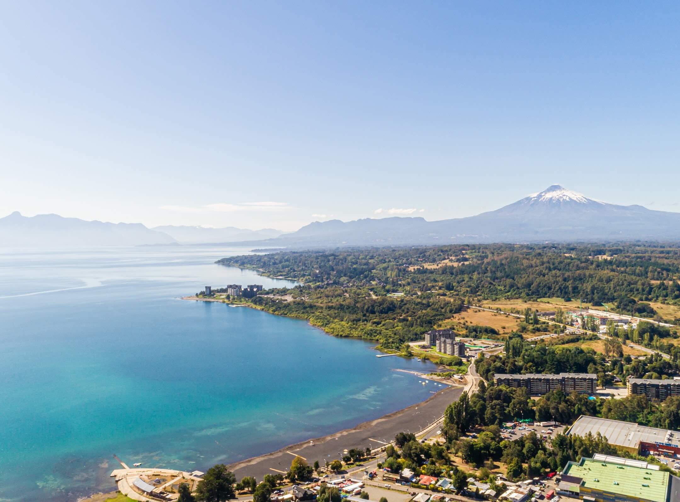 Aerial view of a lake with turquoise water, a shoreline with buildings and greenery, and a snow-capped mountain in the background under a clear blue sky.
