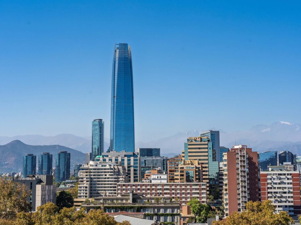 Santiago city skyline with modern high-rise buildings, including a tall glass skyscraper, against a clear blue sky and distant mountains in the background.