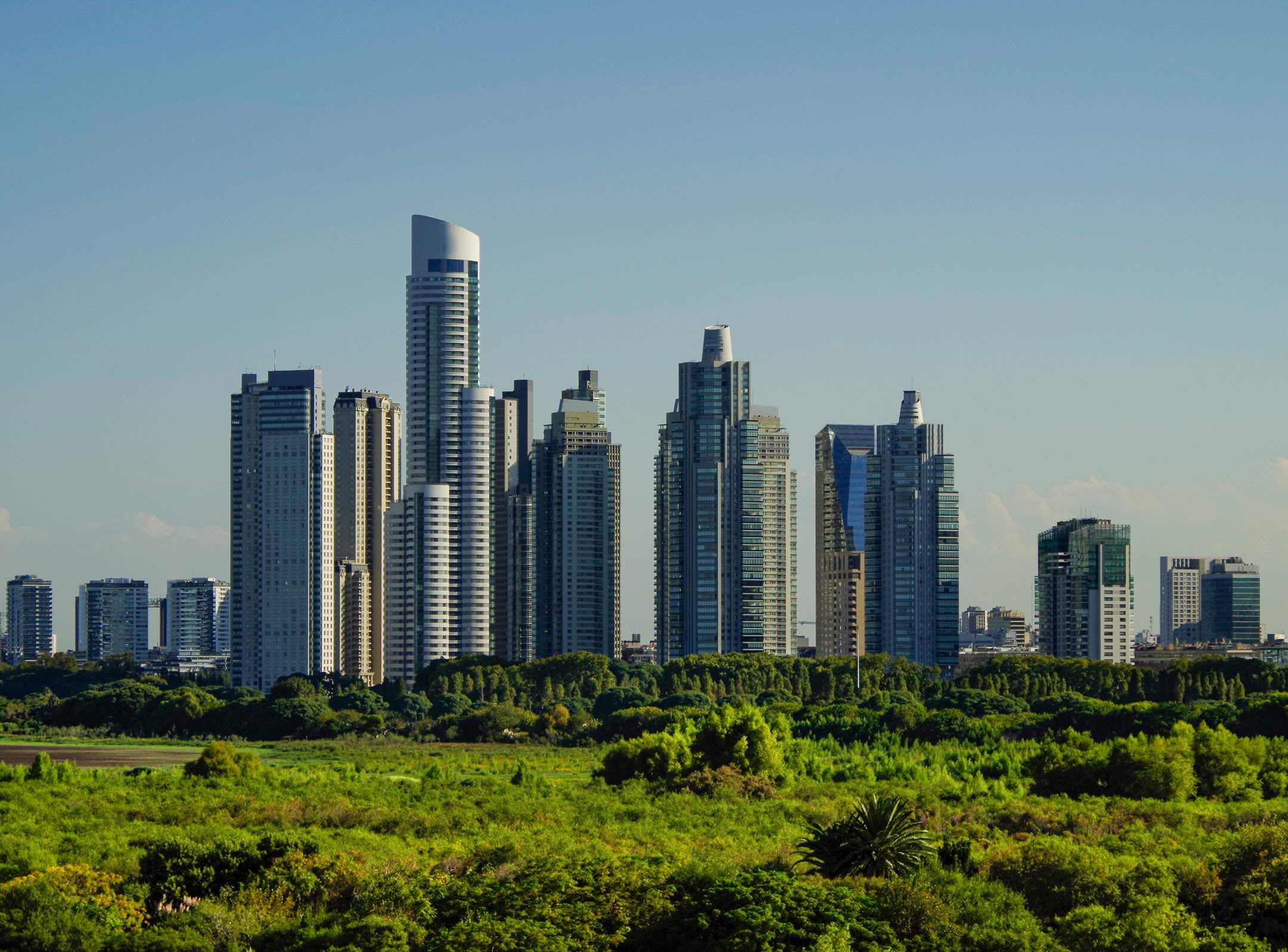 Modern city skyline with tall glass skyscrapers rises above a lush green park under a clear blue sky.