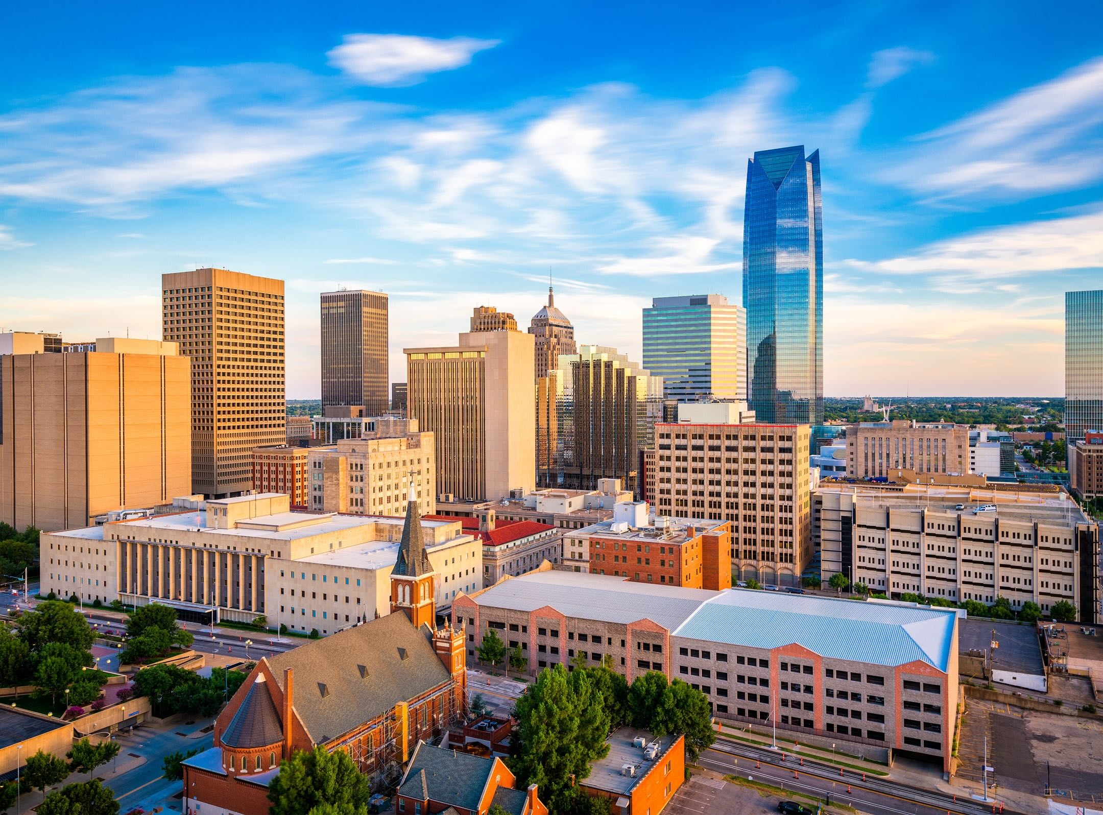 Downtown cityscape featuring modern skyscrapers, mid-rise buildings, and a historic church in the foreground under a blue sky with streaks of clouds.