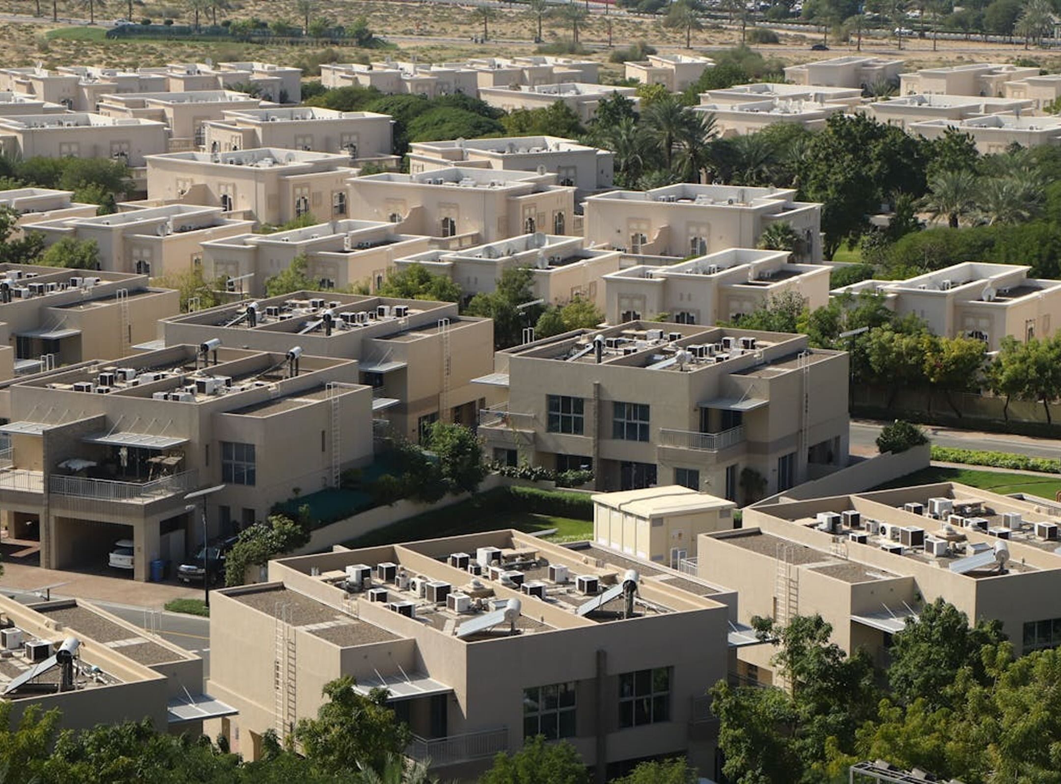 Rows of beige, flat-roofed modern houses with rooftop equipment, surrounded by green trees and vegetation, seen from above in a residential neighborhood.