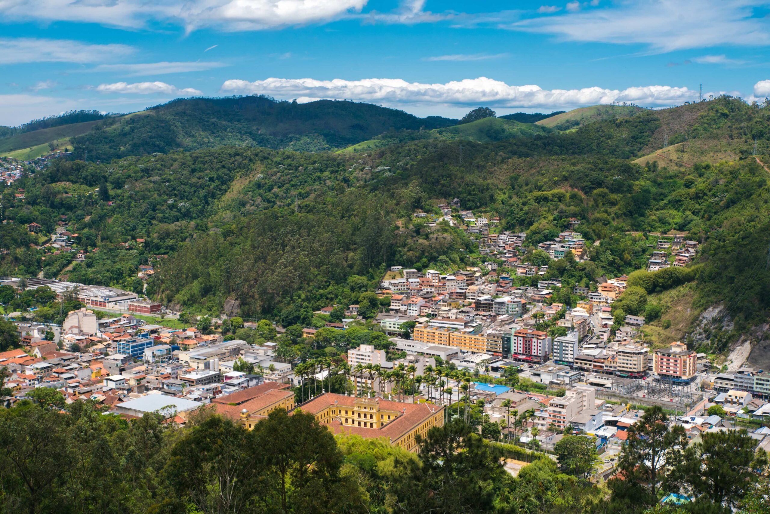 A hillside town with colorful buildings surrounded by green, forested mountains under a blue sky with scattered clouds.