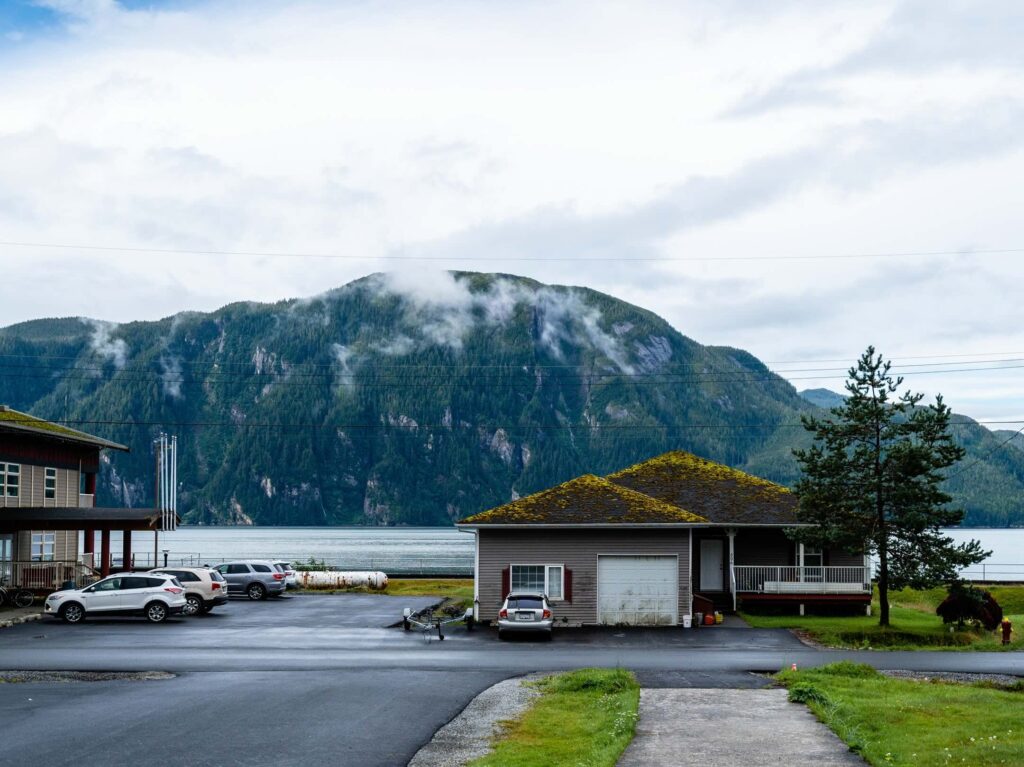 A house with a mossy roof sits near parked cars and a lake, with a large green mountain and low clouds in the background on a cloudy day.
