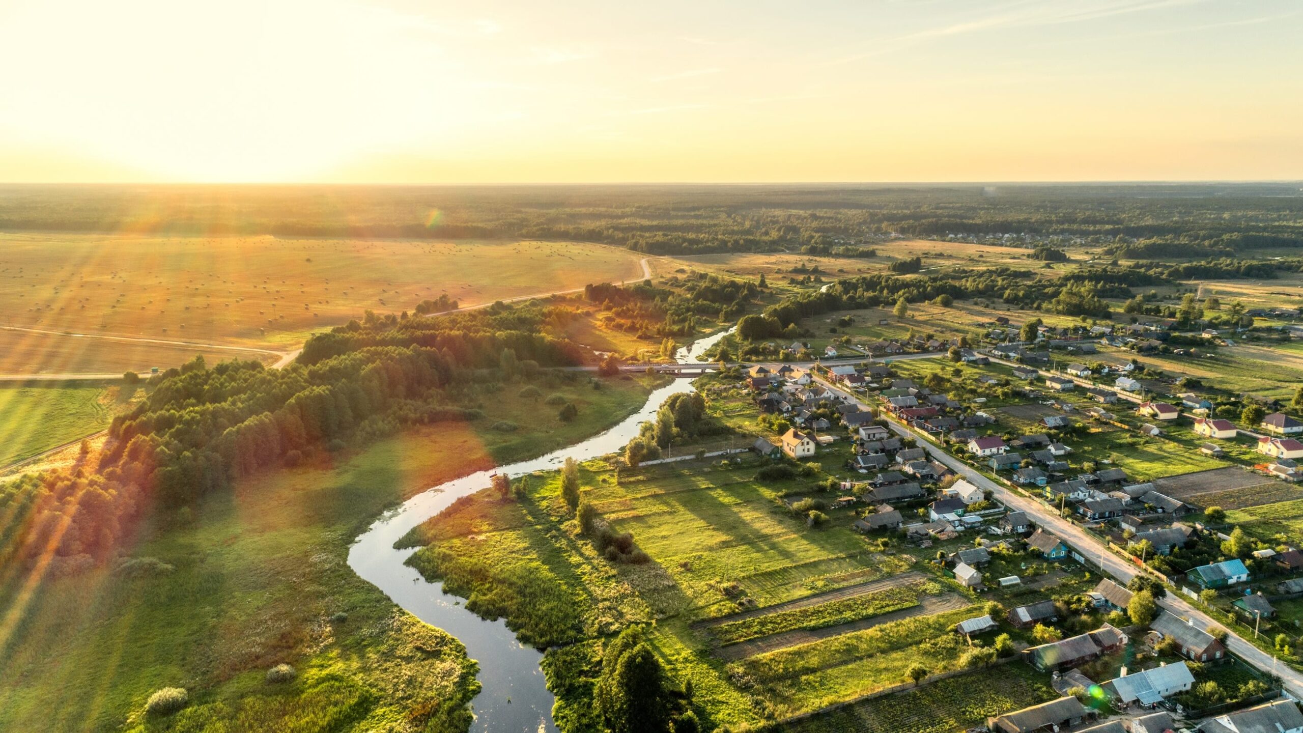 Vista aérea de un pequeño pueblo rural al atardecer, con un río sinuoso, campos verdes, casas y los rayos del sol extendiéndose por el paisaje.