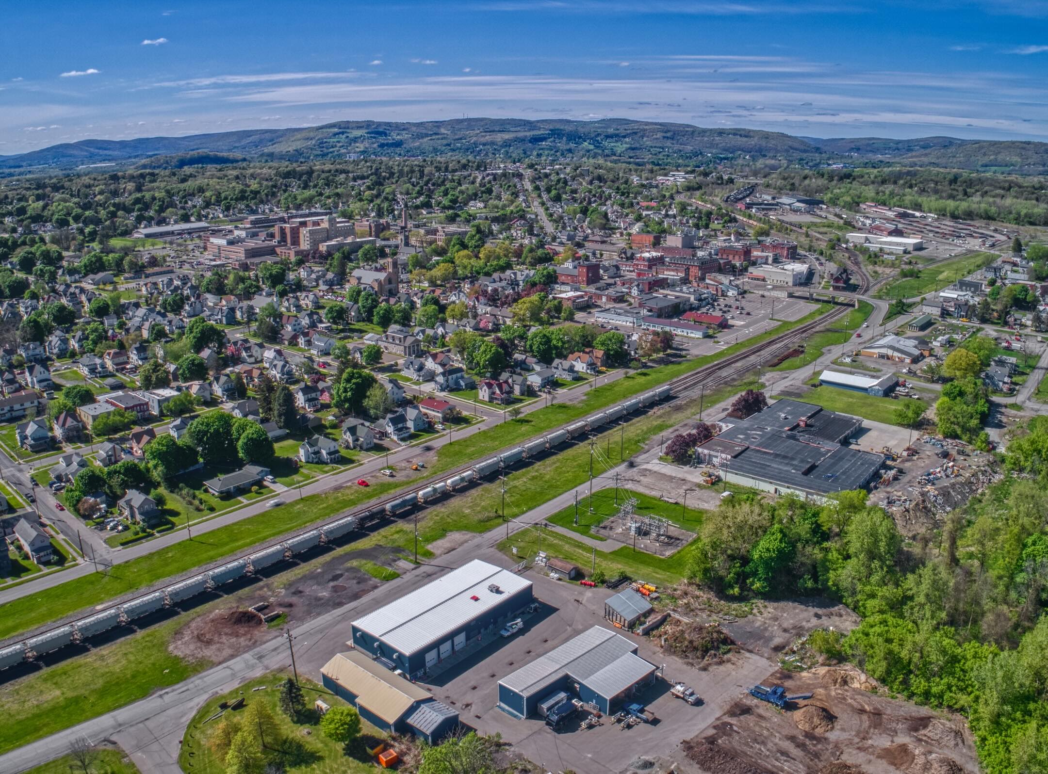 Aerial view of a small town with residential and commercial buildings, a railroad, factories, and green trees under a blue sky with hills in the background.
