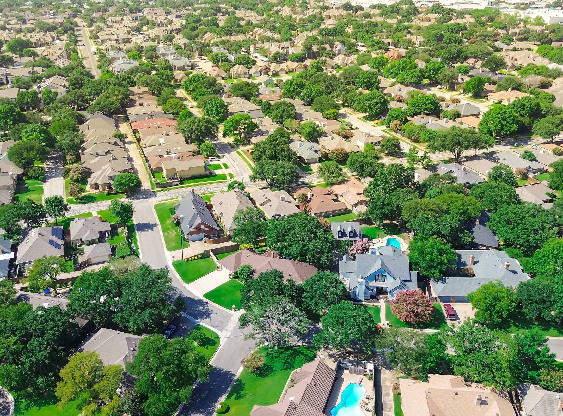 Aerial view of a suburban neighborhood with many houses, green lawns, trees, and visible swimming pools. Streets curve through the area, and rooftops are mostly brown or gray. The scene is bright and sunny.