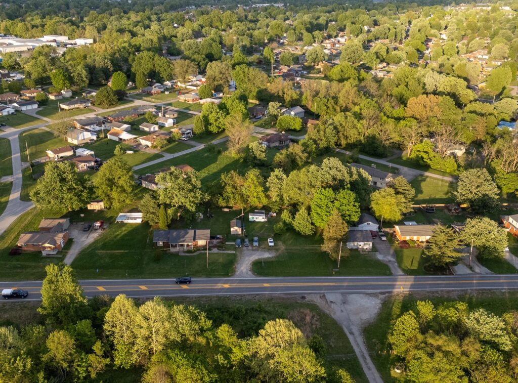 Aerial view of a suburban neighborhood with houses, trees, lawns, and a main road running horizontally in the foreground. The area is lush and green with dense foliage throughout.
