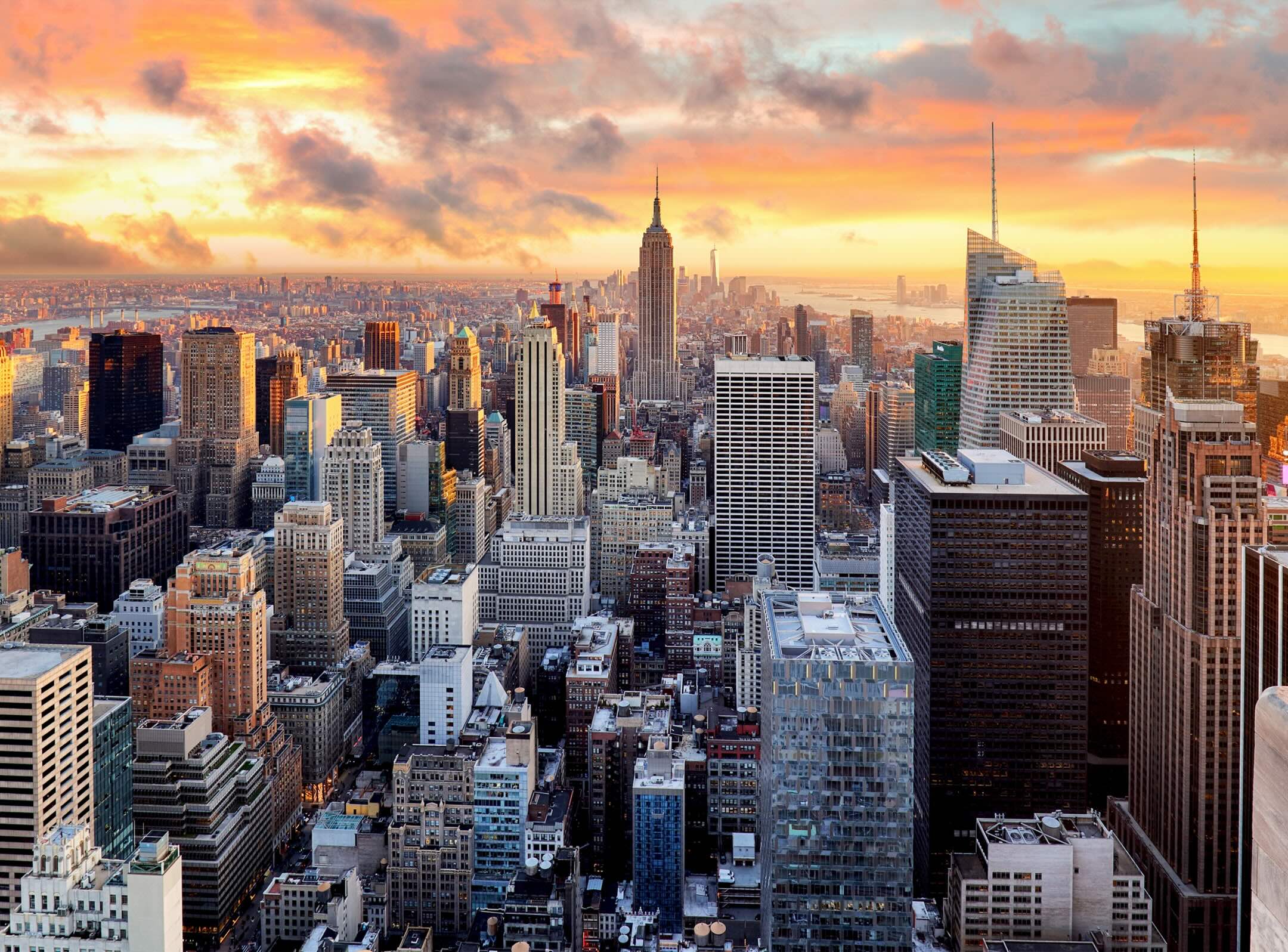 Aerial view of a city skyline at sunset with tall skyscrapers, including a prominent tower in the center, under a colorful sky.