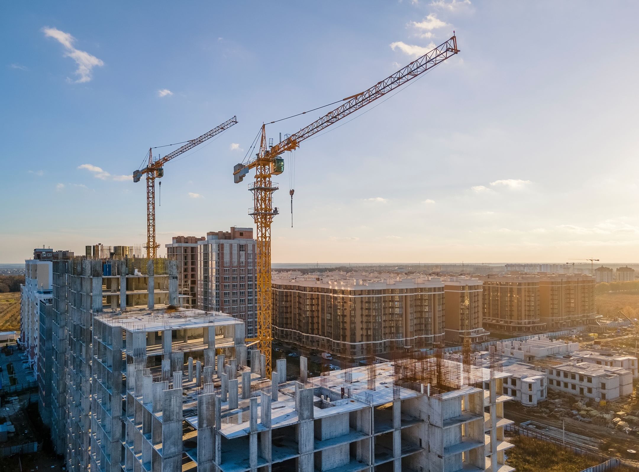 Two large cranes stand over a construction site with partially built buildings in the foreground and several finished apartment complexes in the background under a clear sky.