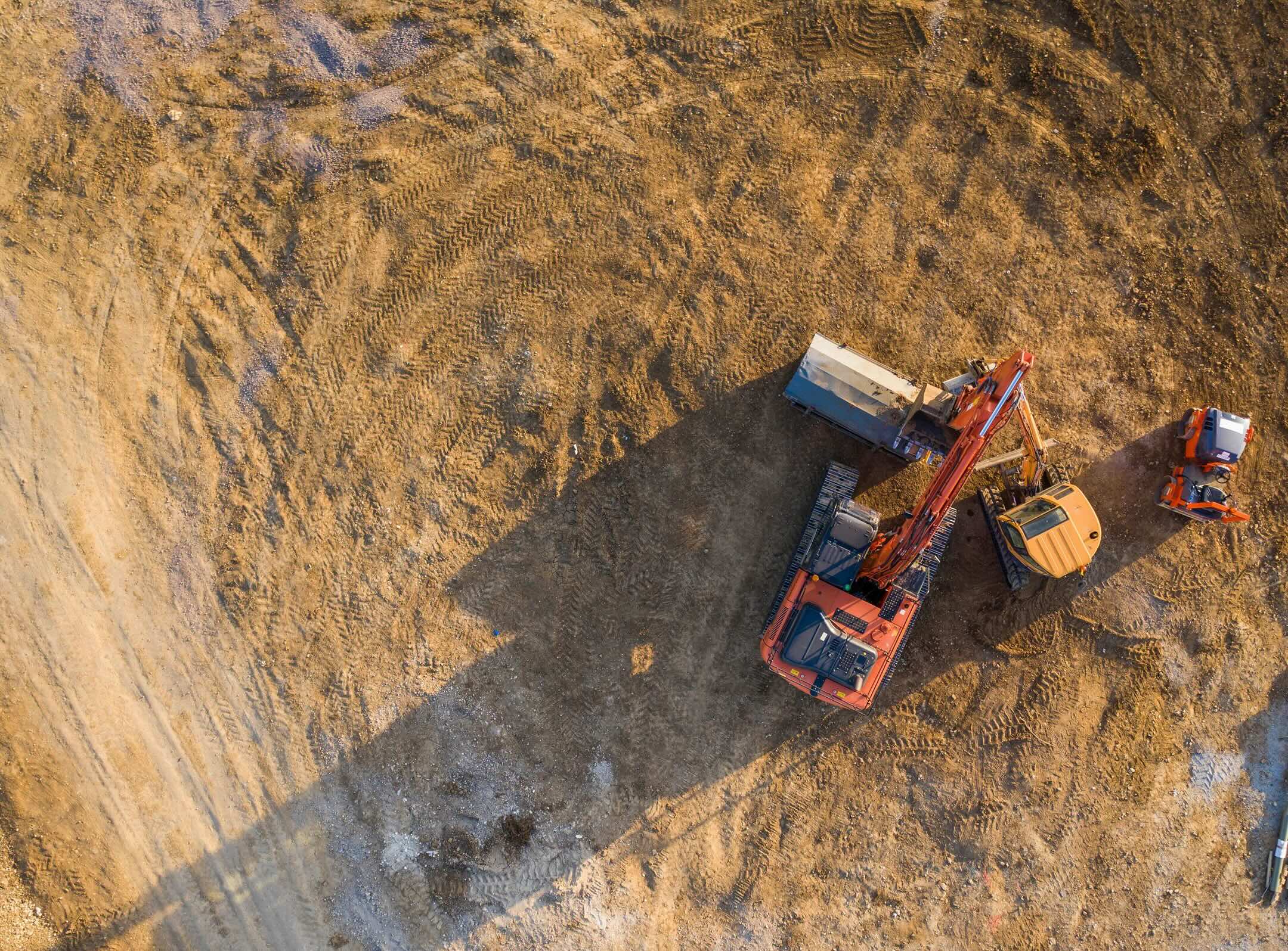 Aerial view of construction vehicles, including an excavator and two trucks, working on a large dirt site with visible tire tracks and machinery shadows.