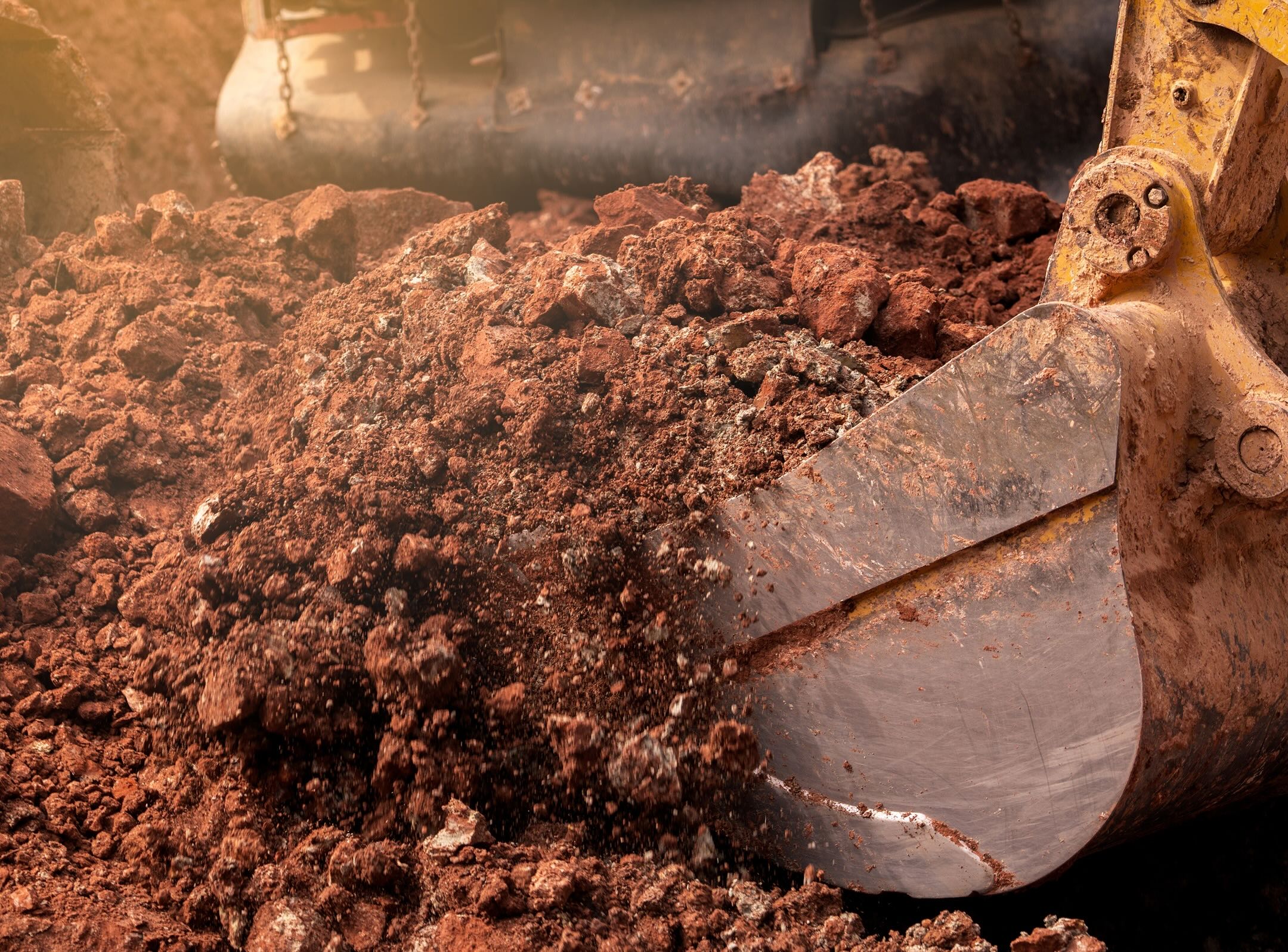 A close-up of an excavator’s metal bucket digging into reddish-brown soil, with loose dirt flying and machinery visible in the background.