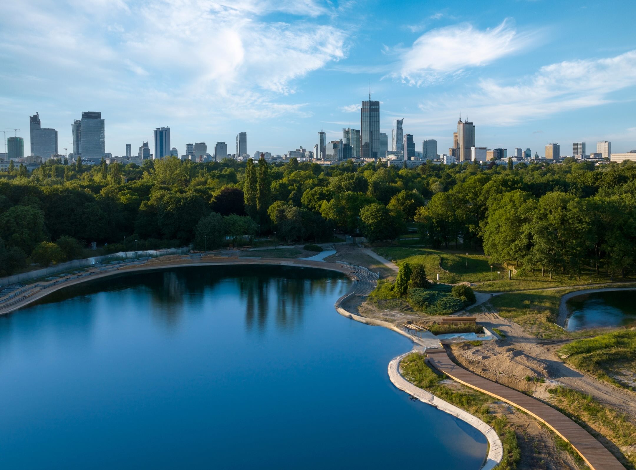A city skyline with tall buildings is seen beyond a large, calm pond bordered by lush green trees and winding paths under a blue sky with wispy clouds.