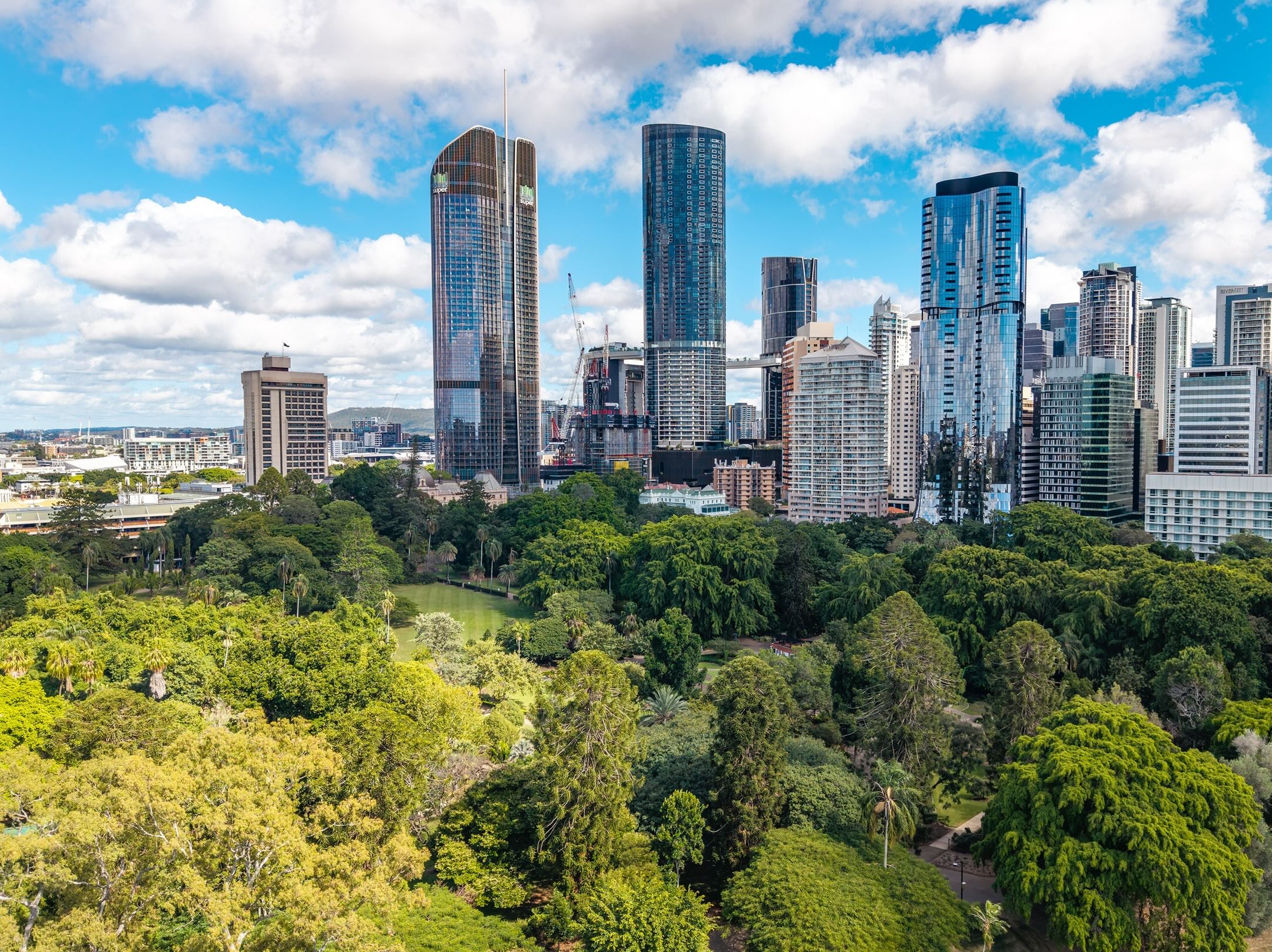 A city skyline with tall modern skyscrapers rises behind a lush, green park filled with trees under a partly cloudy sky.