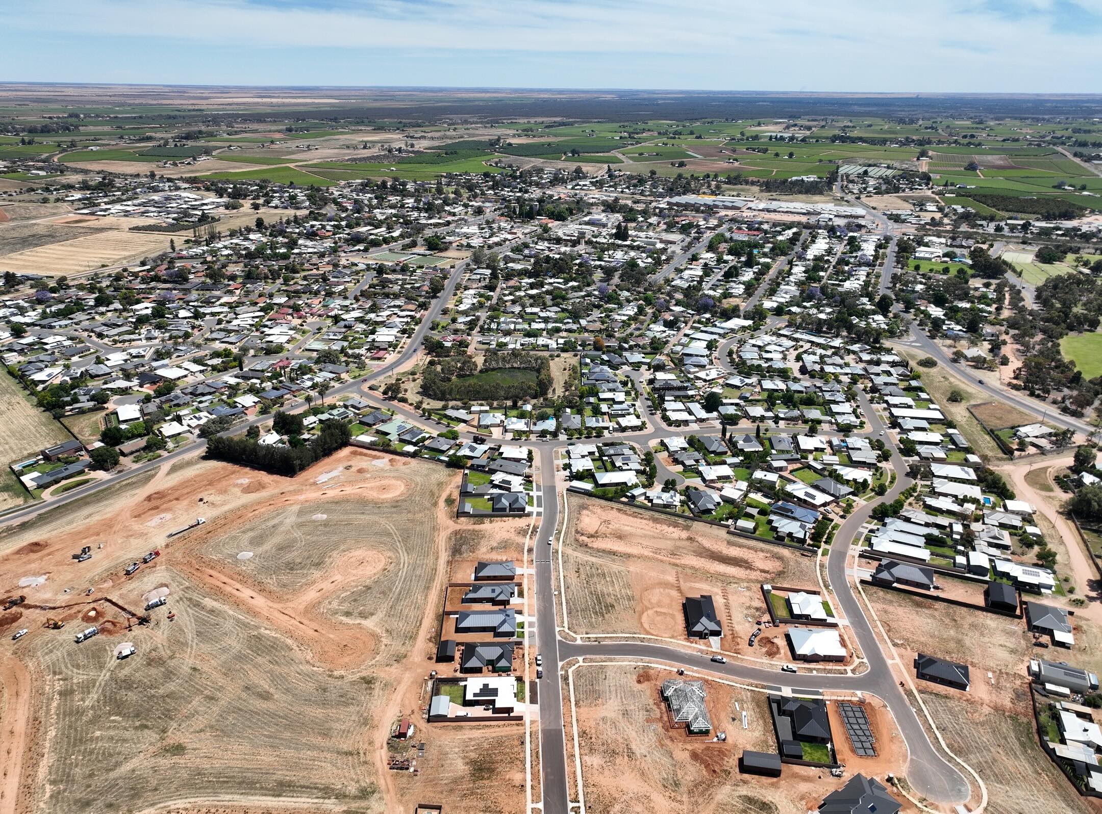 Aerial view of a suburban neighborhood with houses, roads, and open fields. Some areas under construction, surrounding green farmland, and a small town visible in the distance. Blue sky overhead.