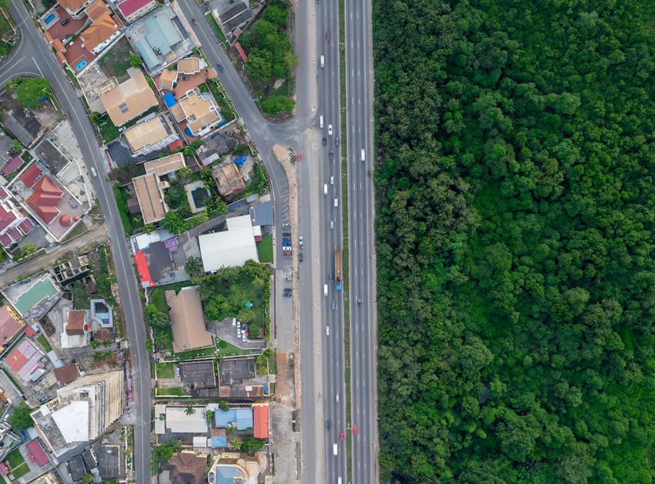 Aerial view of a highway dividing a dense green forest on the right from a residential urban area on the left. Cars are visible on the highway, and rooftops and roads are seen in the neighborhood.