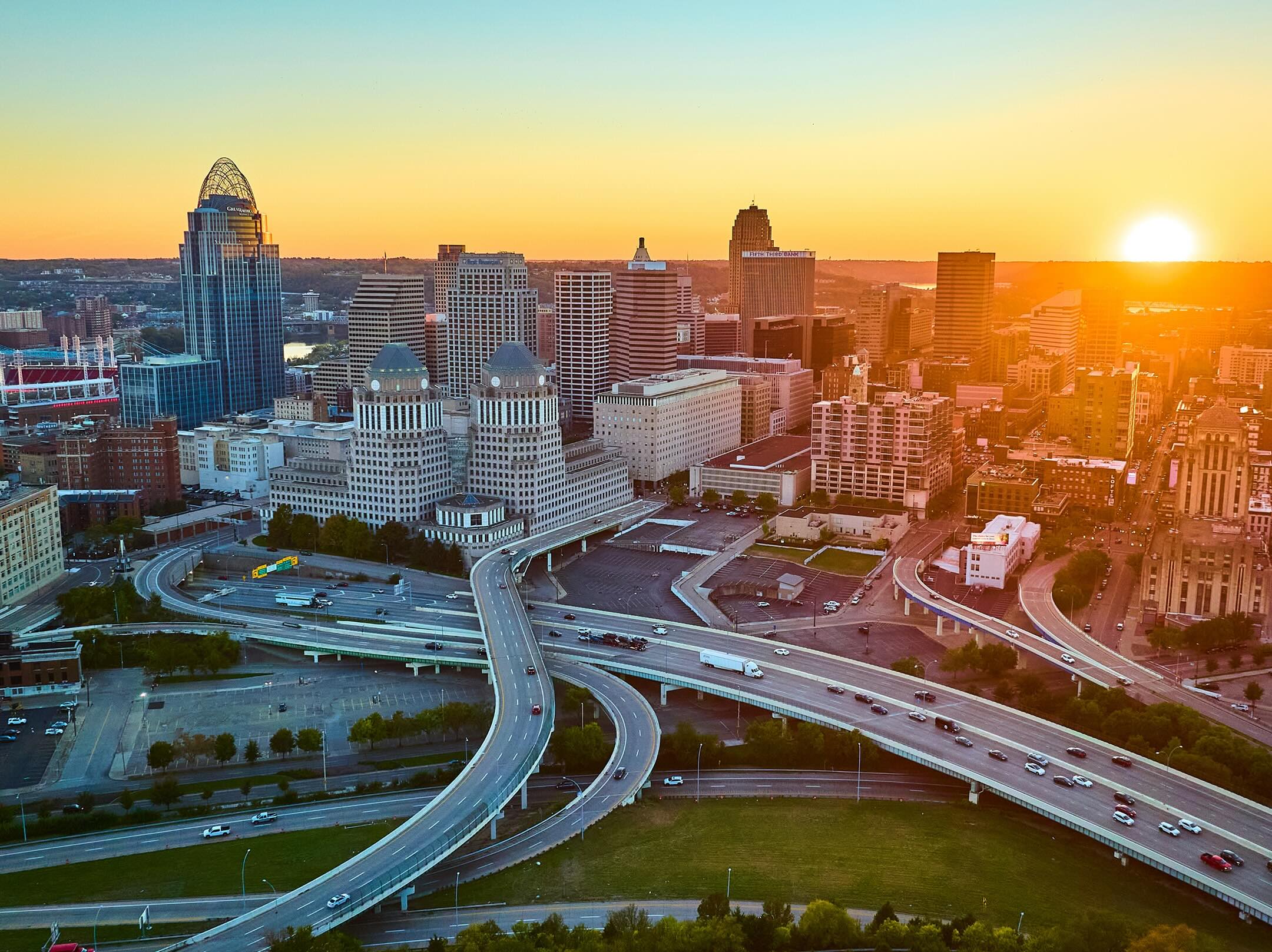 Aerial view of a city skyline at sunset with highways and overpasses in the foreground, tall modern buildings in the background, and the sun setting on the horizon.