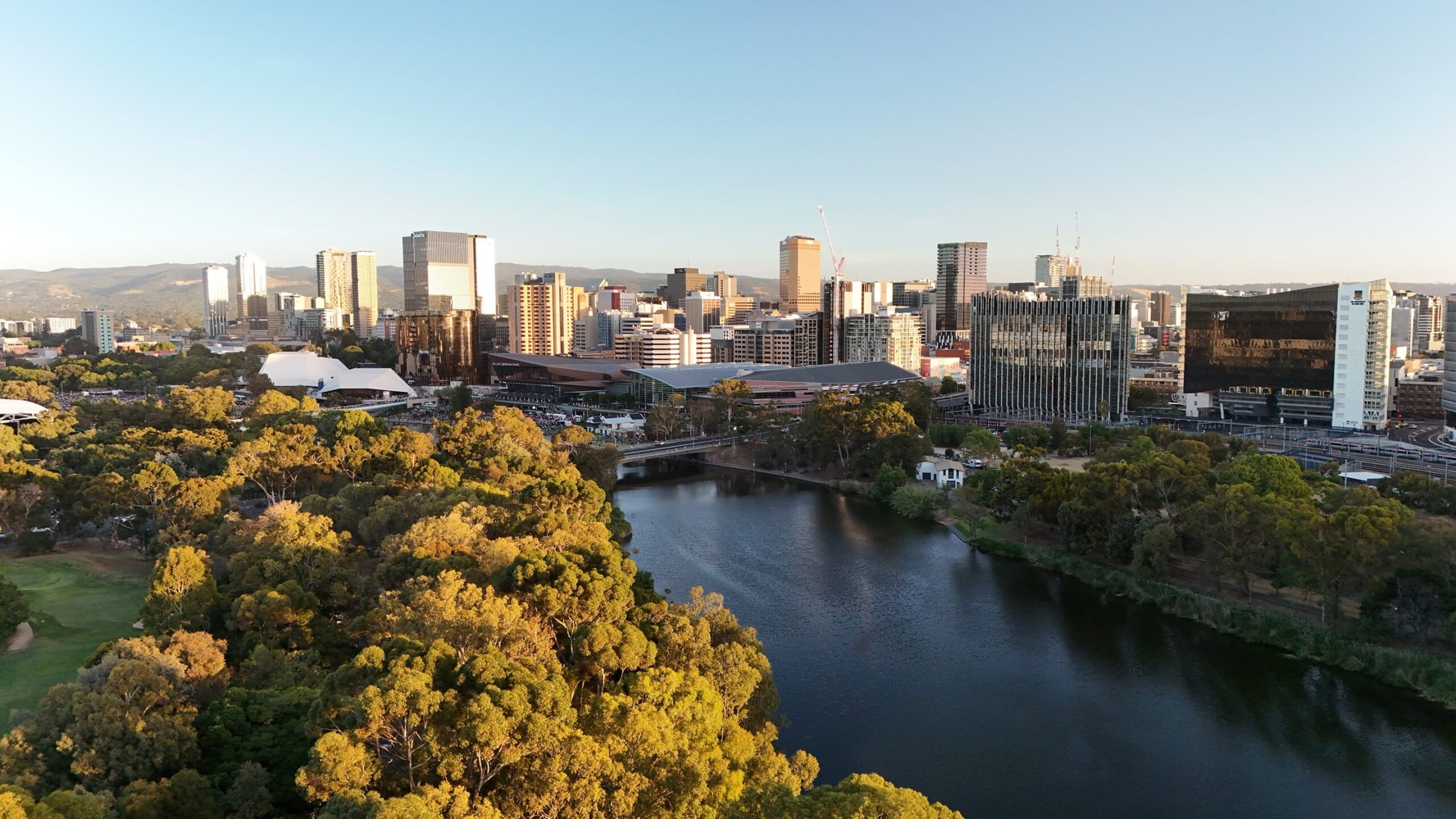 A city skyline with tall buildings, a river running through the foreground, and parks and trees along the water under a clear sky.