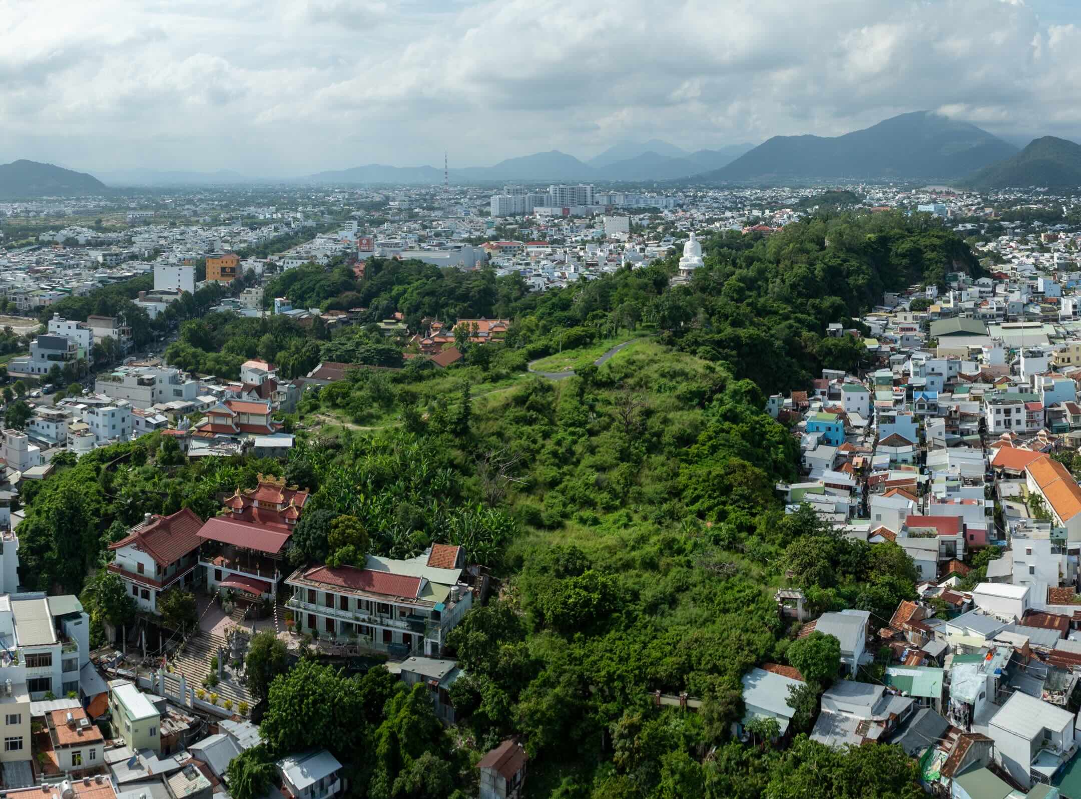 Aerial view of a city with densely packed buildings surrounding a green hill that features a white statue on top, set against distant mountains and a cloudy sky.