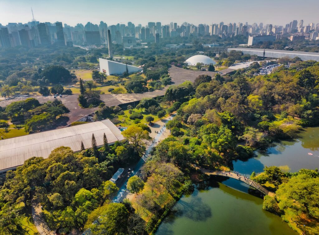Aerial view of Ibirapuera Park in São Paulo, Brazil, showing lush green trees, walking paths, ponds, modern buildings, and the city skyline in the background under a clear sky.