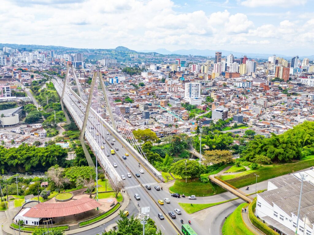 A large cable-stayed bridge crosses a busy city with many cars below, surrounded by dense buildings, greenery, and distant mountains under a partly cloudy sky.