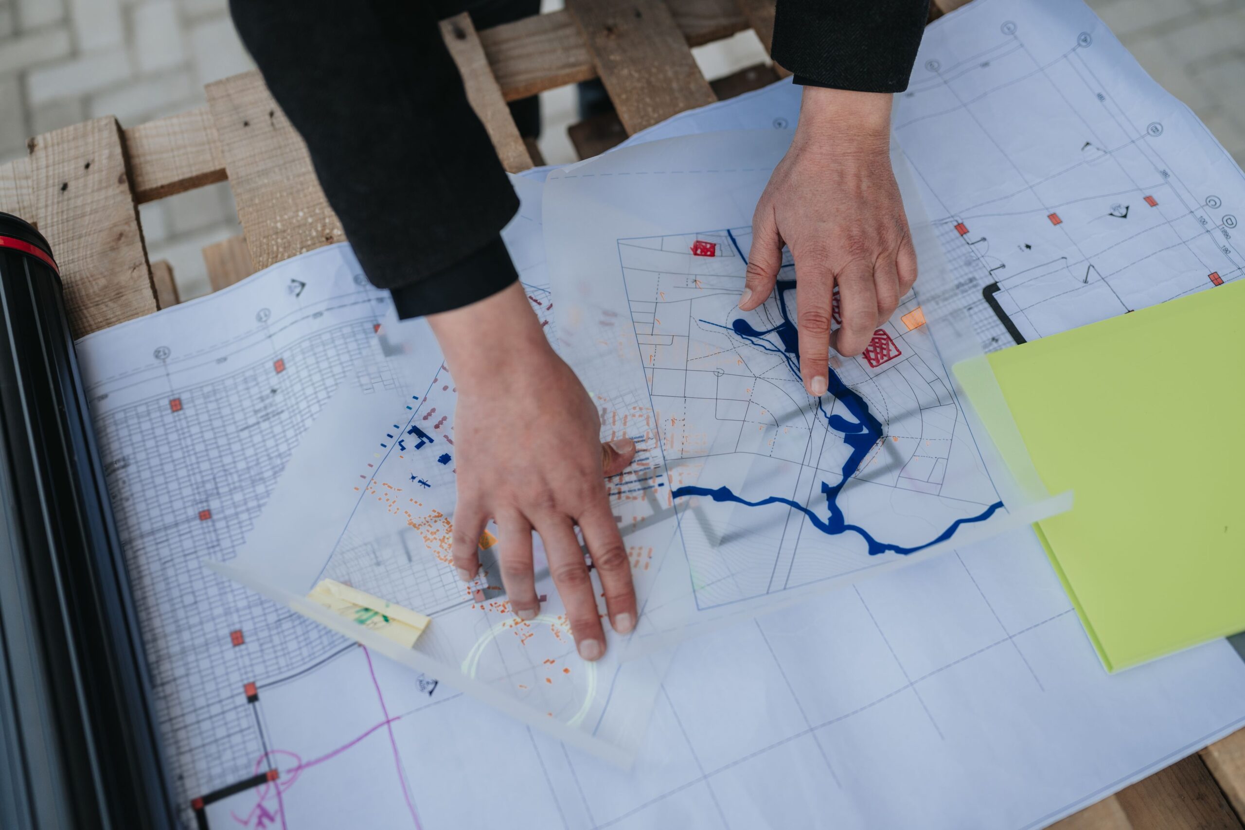 Hands pointing at architectural blueprints and transparent sheets with markings, spread out on a wooden surface alongside a yellow folder and a black tube.