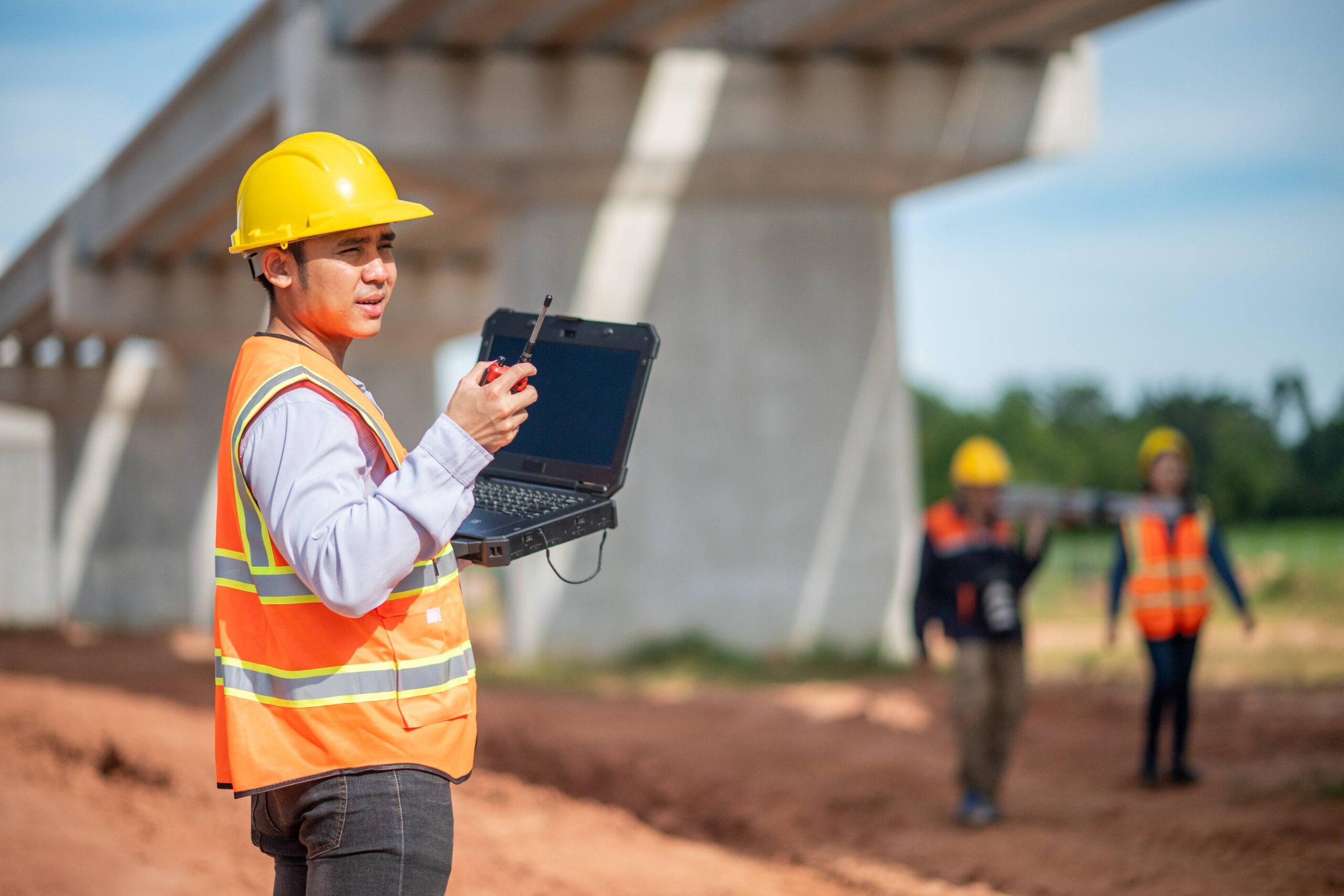 A construction worker in a yellow hard hat and orange vest holds a laptop and walkie-talkie at a construction site, with two other workers in the background near a large concrete structure.
