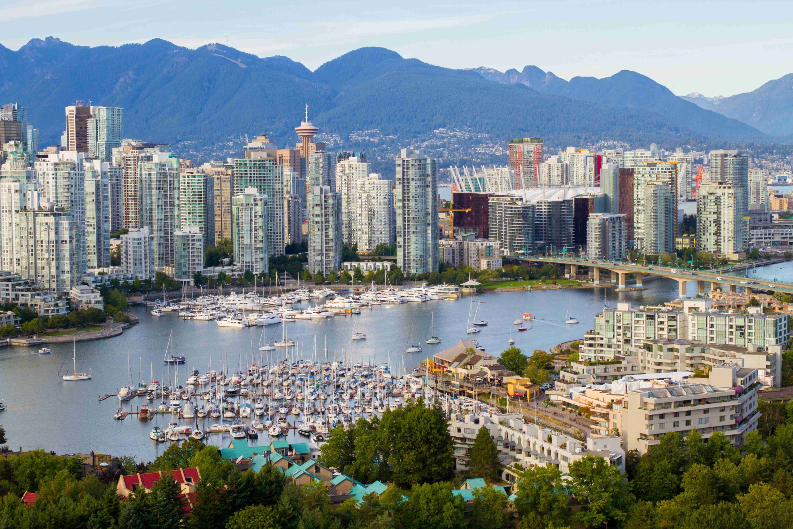 Aerial view of Vancouver, showing downtown skyscrapers, a marina with many boats, a bridge, and mountains in the background on a clear day.