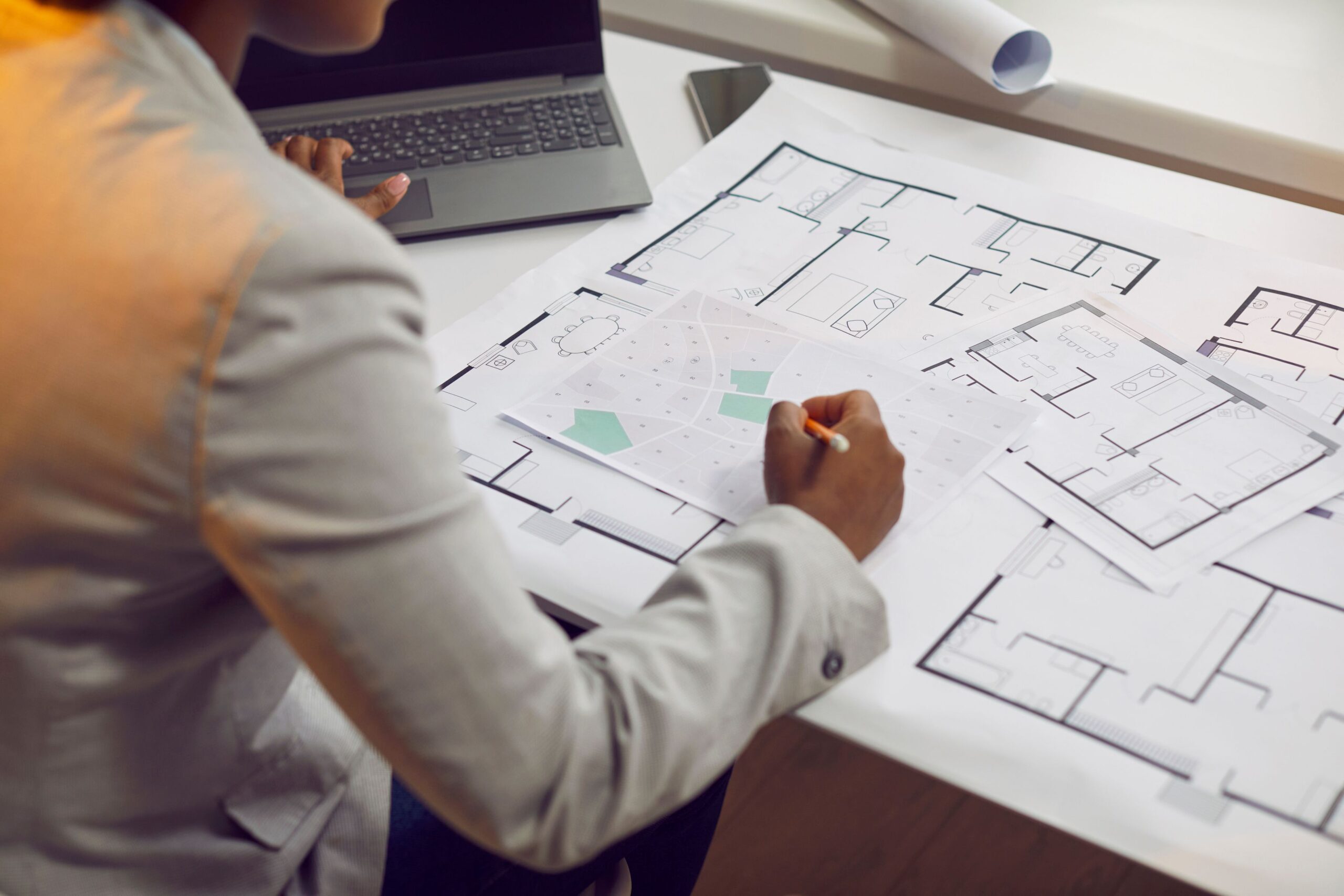 Person in a gray blazer reviews and marks architectural floor plans spread on a desk, with a laptop nearby and rolled-up blueprints in the background.