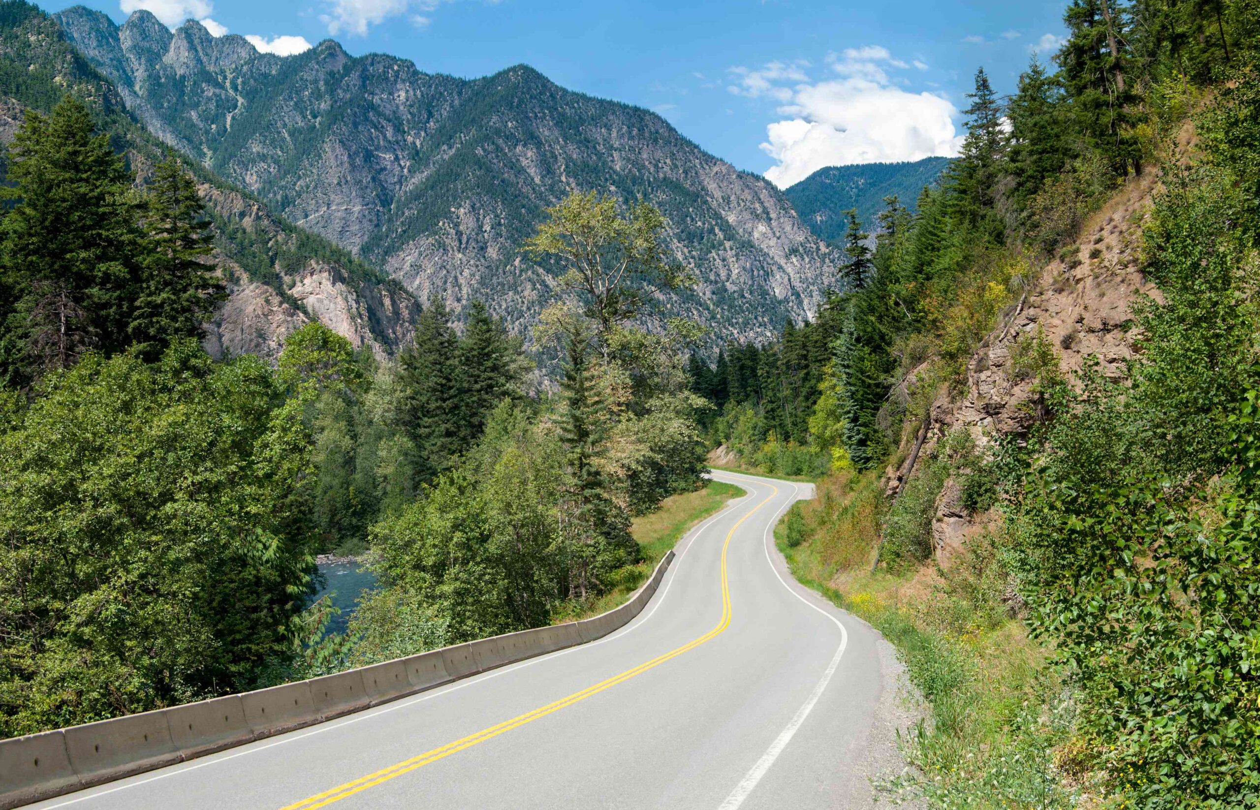 A winding road curves through a lush green forest, with tall mountains and blue sky in the background.